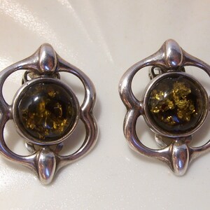 May include: A pair of silver clip-on earrings with a green amber stone in each. The earrings are in the shape of a flower with four petals.