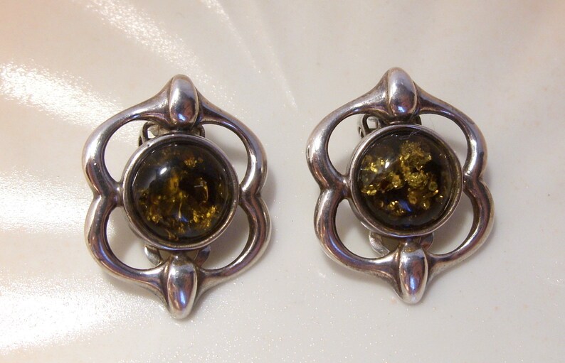 May include: A pair of silver clip-on earrings with a green amber stone in each. The earrings are in the shape of a flower with four petals.