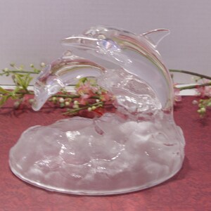 May include: A clear glass sculpture of two dolphins leaping from stylized waves. The dolphins are detailed with fins and facial features. The sculpture is displayed on a red surface with floral accents.