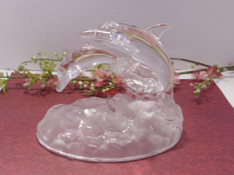 May include: A clear glass sculpture of two dolphins leaping from stylized waves. The dolphins are detailed with fins and facial features. The sculpture is displayed on a red surface with floral accents.