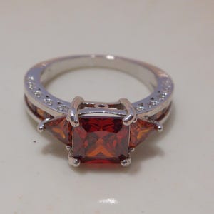 Peut inclure: Bague argentée ornée d'une grosse pierre rouge-orangé carrée, flanquée de deux petites pierres triangulaires. L'anneau présente un motif décoratif. Un bijou.