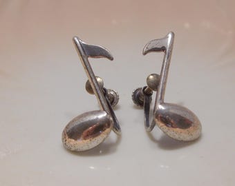 May include: A pair of silver-toned musical note earrings. Each earring features a stylized eighth note design with a curved oval base and a flag. The earrings have a screw-back closure.