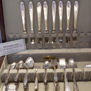 May include: A silver cutlery set presented in a wooden case. The set includes forks, knives, and spoons, neatly arranged within a grey felt-lined interior. The silverware features decorative handles. A card with the text "WM. ROGERS & SON" is visible.