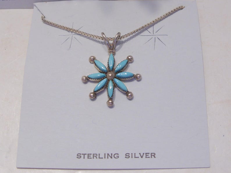 May include: A silver necklace with a turquoise flower pendant. The pendant is made of sterling silver and has a blue turquoise stone in the center. The necklace is on a white background with the words "STERLING SILVER" printed below the pendant.