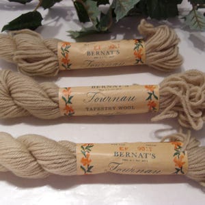 May include: Three skeins of beige Bernat's Tournau Tapestry Wool yarn. Each skein is wrapped with a paper label featuring floral designs and the brand name. The yarn is tightly wound, showing the texture of the wool.