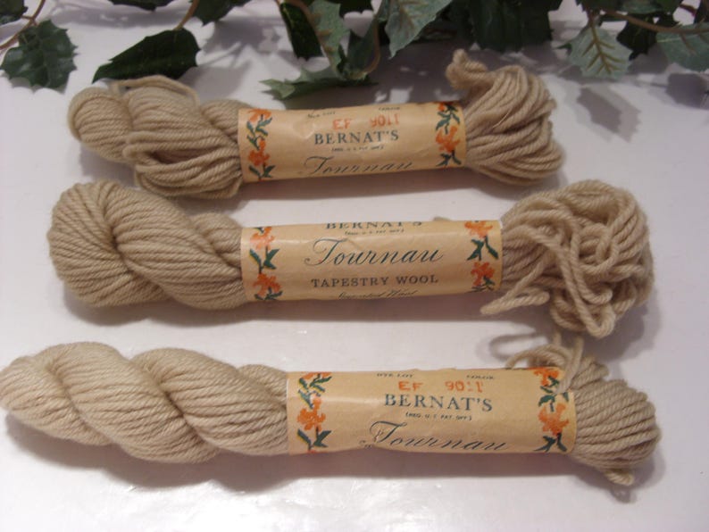 May include: Three skeins of beige Bernat's Tournau Tapestry Wool yarn. Each skein is wrapped with a paper label featuring floral designs and the brand name. The yarn is tightly wound, showing the texture of the wool.