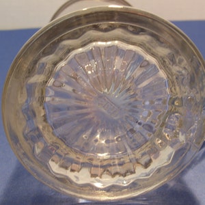 May include: A clear glass pitcher with a silver rim and handle. The bottom of the pitcher has a textured, starburst design. The pitcher is sitting on a blue surface.