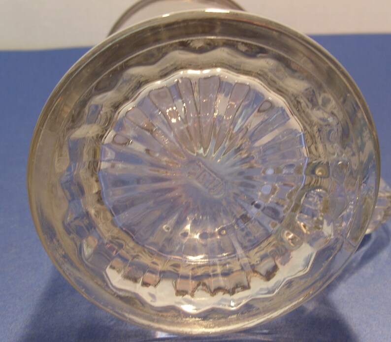 May include: A clear glass pitcher with a silver rim and handle. The bottom of the pitcher has a textured, starburst design. The pitcher is sitting on a blue surface.