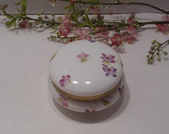 May include: A white porcelain trinket box with a floral design. The lid features a delicate pattern of pink and purple flowers. The box has a gold trim around the edge.