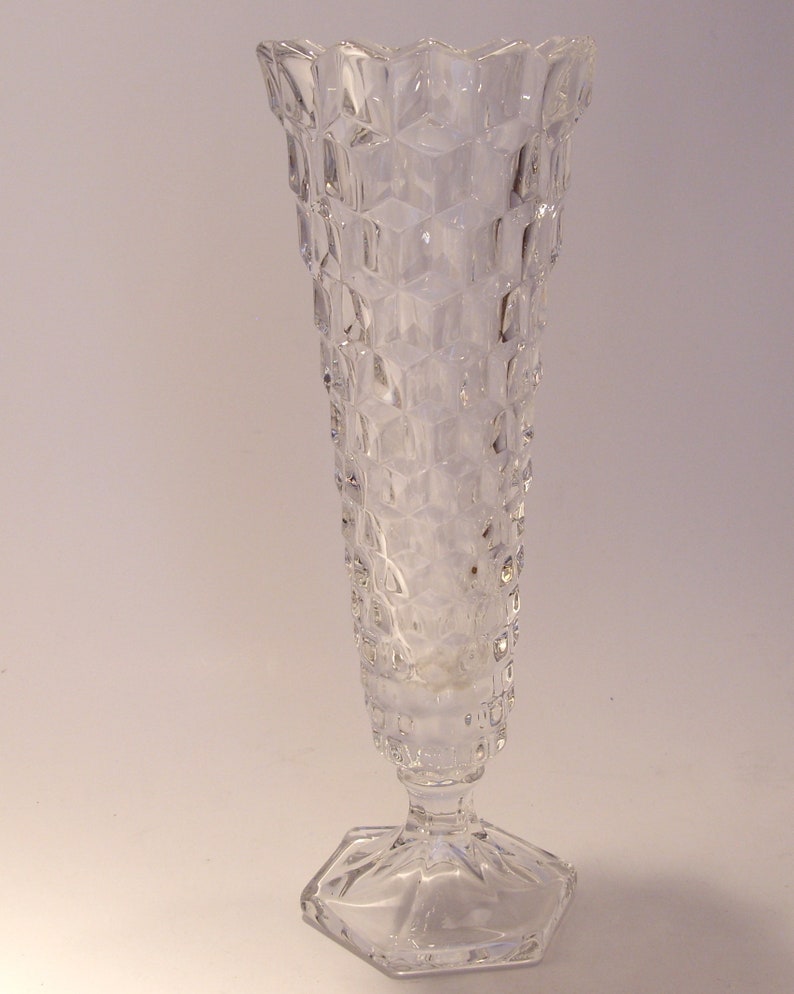 Puede incluir: Jarr&oacute;n de cristal transparente con un patr&oacute;n geom&eacute;trico texturizado. El jarr&oacute;n tiene una base hexagonal y una parte superior acampanada y dentada. El cuerpo del jarr&oacute;n es largo y esbelto.