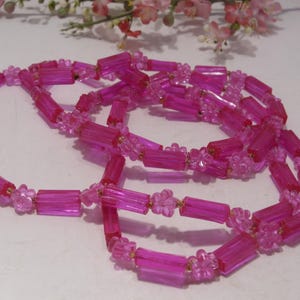 May include: A long necklace composed of translucent hot pink cylindrical beads alternating with small, clear pink flower-shaped beads. The strand is arranged in a loose, coiled loop, showcasing the repeating pattern of the bright pink plastic components.