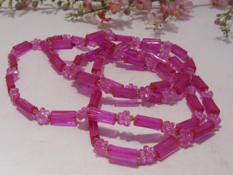 May include: A long necklace composed of translucent hot pink cylindrical beads alternating with small, clear pink flower-shaped beads. The strand is arranged in a loose, coiled loop, showcasing the repeating pattern of the bright pink plastic components.