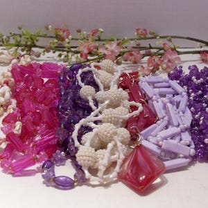 May include: A collection of six necklaces featuring various styles, including white seashell strands, translucent hot pink rectangular beads, deep purple faceted beads, white textured spherical beads, lavender rectangular beads, and purple seed beads.