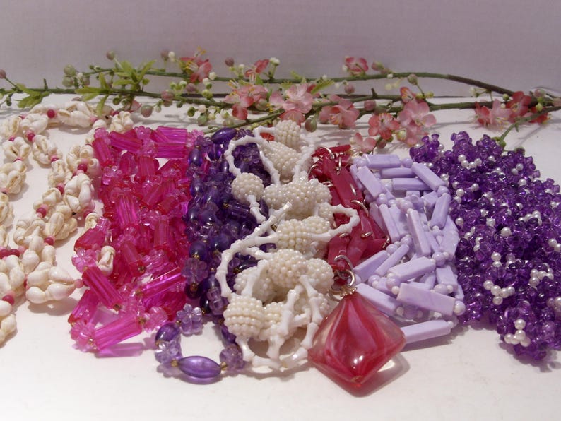 May include: A collection of six necklaces featuring various styles, including white seashell strands, translucent hot pink rectangular beads, deep purple faceted beads, white textured spherical beads, lavender rectangular beads, and purple seed beads.