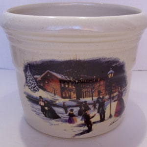 May include: Off-white ceramic planter with a winter scene illustration. The image depicts people ice skating and walking near buildings and a lamppost. The planter has a slightly flared rim and a smooth, untextured surface.