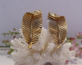 May include: A pair of gold-toned metal earrings shaped like stylized leaves. Each earring features a central raised vein with textured, ribbed details extending outward to the edges, creating a realistic leaf-like appearance with a polished finish.