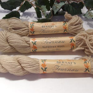 May include: Three skeins of beige Bernat's Tournau Tapestry Wool yarn. Each skein is wrapped with a paper label featuring floral accents and the yarn's name. The yarn is arranged on a white surface, with greenery in the background.