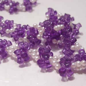 May include: A necklace featuring a repeating pattern of translucent purple, spool-shaped plastic beads interspersed with small, round, white faux pearls. The beads are strung together in a continuous, flexible strand with a textured, clustered appearance.