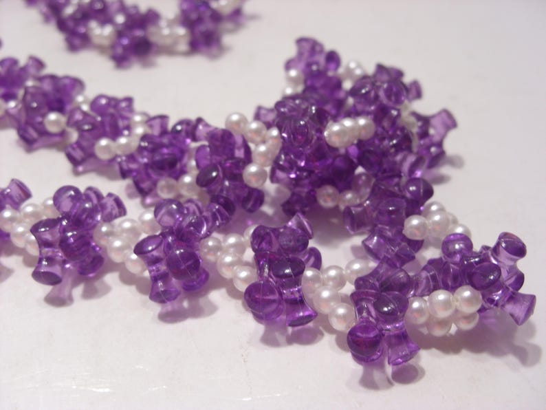 May include: A necklace featuring a repeating pattern of translucent purple, spool-shaped plastic beads interspersed with small, round, white faux pearls. The beads are strung together in a continuous, flexible strand with a textured, clustered appearance.