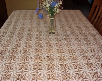 May include: A white crocheted tablecloth with a delicate floral pattern covers a wooden table. A vase with blue and white flowers sits in the center of the table.