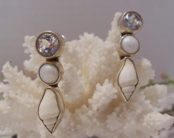 May include: A pair of drop earrings featuring a vertical arrangement of a clear faceted stone, a smooth white round bead, and a small white spiral seashell, all set in a metallic frame with a post back closure for secure attachment.