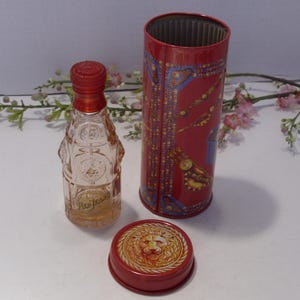May include: A clear glass perfume bottle with a red cap, accompanied by a cylindrical red metal tin featuring gold and blue decorative patterns. A separate red metal lid displays a raised lion head emblem in the center of the circular design.