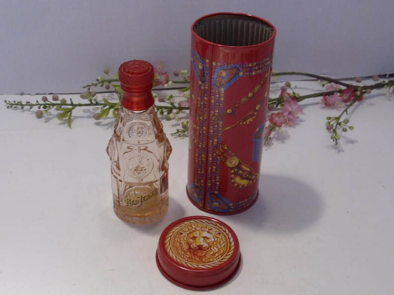 May include: A clear glass perfume bottle with a red cap, accompanied by a cylindrical red metal tin featuring gold and blue decorative patterns. A separate red metal lid displays a raised lion head emblem in the center of the circular design.