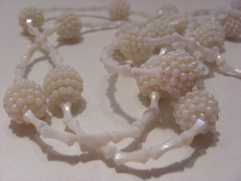 May include: A necklace featuring a series of white, textured, spherical beads alternating with smooth, elongated, bone-shaped spacers. The beads have a glossy, pearlescent finish, and the entire piece is constructed with a repeating pattern.