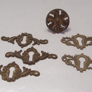 May include: Antique bronze-colored hardware set, including a round knob and five ornate keyhole escutcheons. The decorative pieces feature intricate floral and scrollwork designs, ideal for vintage furniture restoration projects.