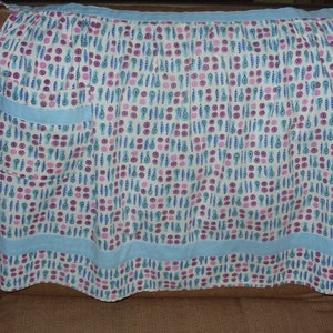 May include: A white apron with a blue trim and a pocket. The apron has a pattern of pink, blue, and green pears and circles.