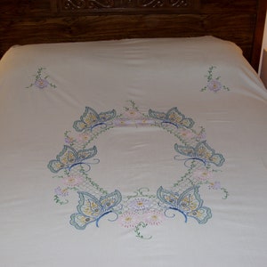 May include: Cream-colored bedspread with embroidered butterflies and floral accents. The central design features a wreath of butterflies and flowers, with additional floral clusters in the corners. The butterflies have blue and yellow details.