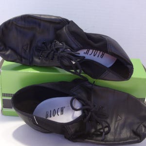 Puede incluir: Un par de zapatos de baile de cuero negro con cordones, apoyados en una caja verde. Los zapatos tienen un dise&ntilde;o suave y flexible y la marca "BLOCH" es visible en la plantilla. Zapatos de baile.