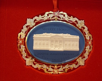 May include: A Christmas ornament featuring a white relief of the White House on a blue oval. The oval is framed by a gold and white filigree design. Text reads "The White House 200th Anniversary Christmas 2000".