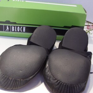 Puede incluir: Par de zapatillas de ballet negras con suelas texturizadas, delante de una caja verde con la marca "BLOCH". Las zapatillas tienen una puntera redondeada y un dise&ntilde;o suave y flexible, adecuado para la pr&aacute;ctica de la danza.