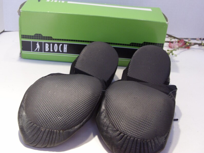 Puede incluir: Par de zapatillas de ballet negras con suelas texturizadas, delante de una caja verde con la marca "BLOCH". Las zapatillas tienen una puntera redondeada y un dise&ntilde;o suave y flexible, adecuado para la pr&aacute;ctica de la danza.