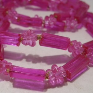 May include: A necklace featuring translucent hot pink beads in an alternating pattern of elongated rectangular tubes and small five-petaled flowers. The beads are strung together on a thin cord, creating a continuous loop of bright pink plastic components.