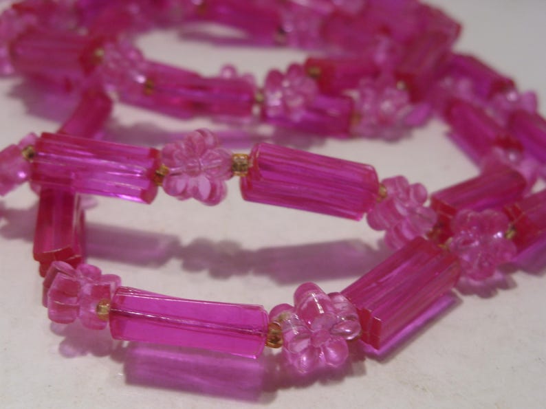 May include: A necklace featuring translucent hot pink beads in an alternating pattern of elongated rectangular tubes and small five-petaled flowers. The beads are strung together on a thin cord, creating a continuous loop of bright pink plastic components.