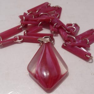 May include: A necklace featuring a series of rectangular, dark pink, marbled links connected by silver-toned metal rings. The central pendant is a diamond-shaped, clear acrylic piece with a swirling, dark pink and white marbled core.