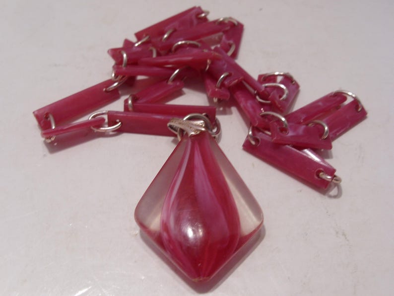 May include: A necklace featuring a series of rectangular, dark pink, marbled links connected by silver-toned metal rings. The central pendant is a diamond-shaped, clear acrylic piece with a swirling, dark pink and white marbled core.