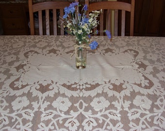 May include: A white lace tablecloth with a delicate floral pattern. A glass vase with blue and white flowers sits in the center of the table.
