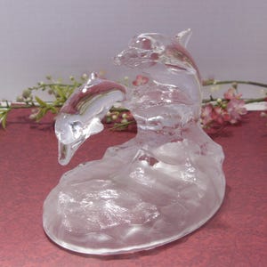May include: A clear glass dolphin sculpture with a mother and baby dolphin leaping through a wave. The sculpture has a frosted appearance and sits on a burgundy surface. Decorative flowers are in the background.