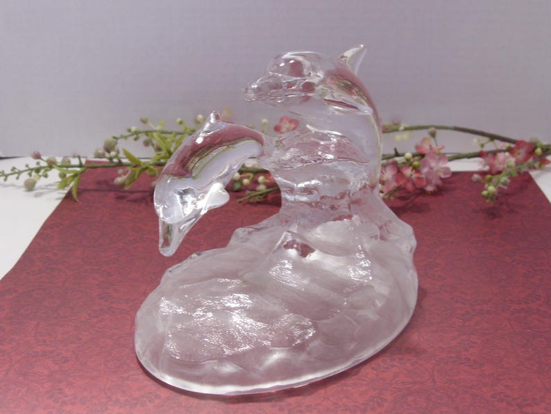 May include: A clear glass dolphin sculpture with a mother and baby dolphin leaping through a wave. The sculpture has a frosted appearance and sits on a burgundy surface. Decorative flowers are in the background.