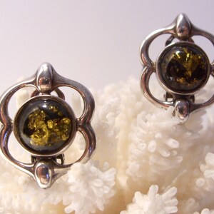 May include: A pair of silver clip-on earrings with a green amber stone in the center of each earring. The earrings are shaped like a flower with four petals.