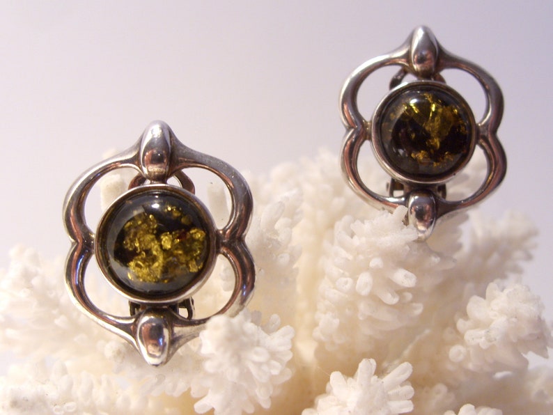 May include: A pair of silver clip-on earrings with a green amber stone in the center of each earring. The earrings are shaped like a flower with four petals.