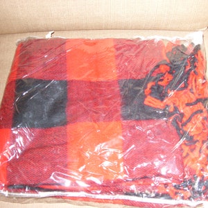 Op de afbeelding: Een rood-zwarte geruite deken, verpakt in doorzichtig plastic. De deken heeft een klassiek buffelruitpatroon. De franje is zichtbaar aan de rand. De deken is opgevouwen en klaar voor gebruik.