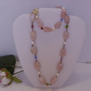 May include: A long necklace featuring large, light pink, oval-shaped beads. The necklace is accented with small, colorful beads and pearls. It is displayed on a white bust form, with a gold-colored clasp.