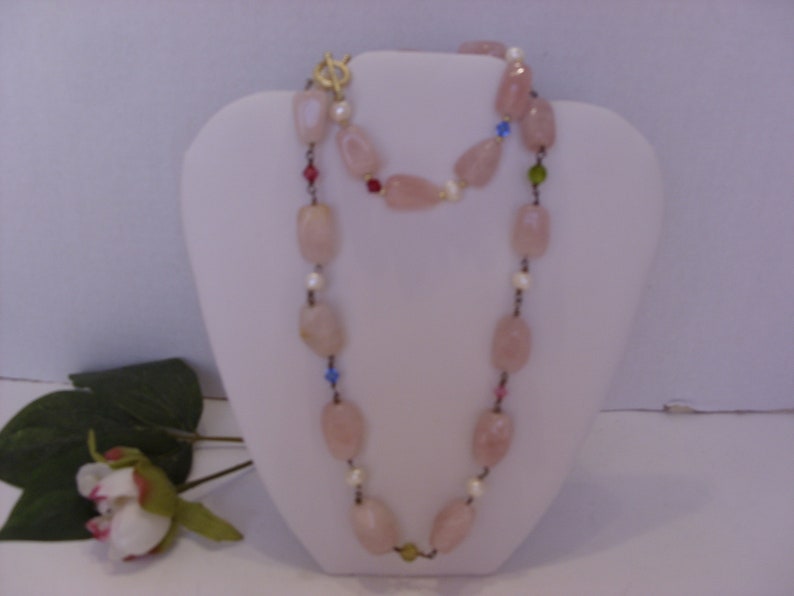 May include: A long necklace featuring large, light pink, oval-shaped beads. The necklace is accented with small, colorful beads and pearls. It is displayed on a white bust form, with a gold-colored clasp.