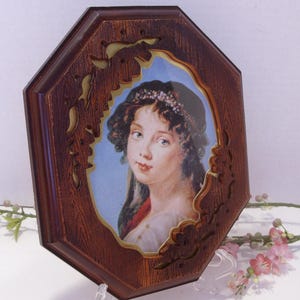 May include: An octagonal wooden framed portrait of a woman with a floral crown. The portrait features a woman with light skin, blue eyes, and dark curly hair. The frame has a dark brown wood grain finish with decorative cutouts.