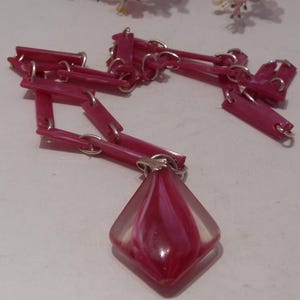 May include: A necklace featuring a chain of rectangular pink plastic links connected by silver-toned metal rings. The pendant is a diamond-shaped, translucent glass piece with a swirling pattern of deep pink and white colors, attached by a silver bail.
