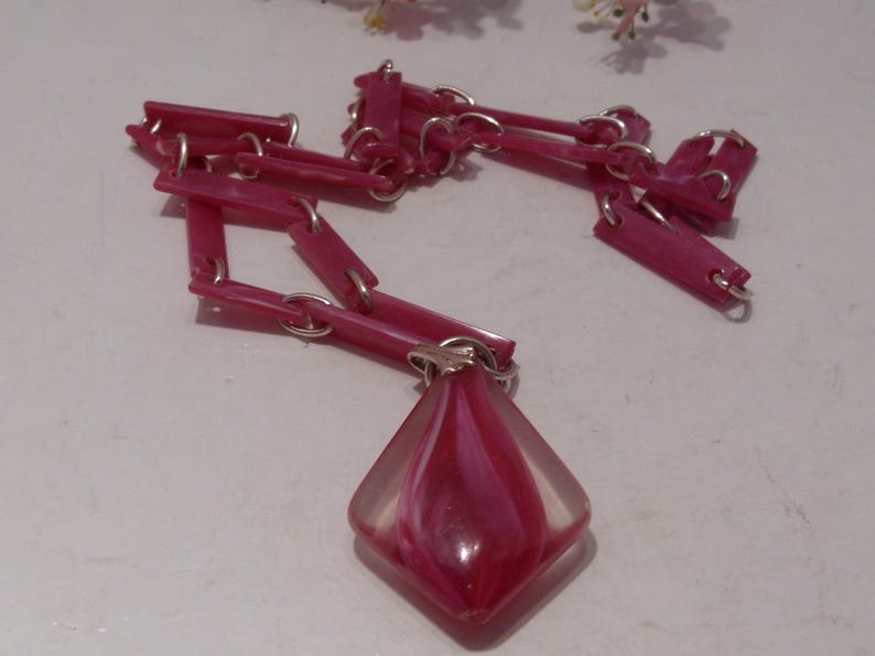 May include: A necklace featuring a chain of rectangular pink plastic links connected by silver-toned metal rings. The pendant is a diamond-shaped, translucent glass piece with a swirling pattern of deep pink and white colors, attached by a silver bail.